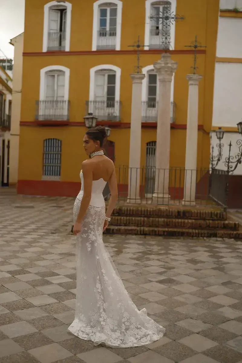 A bride in a white wedding dress with floral details stands elegantly in front of historical pillars and a colorful building.