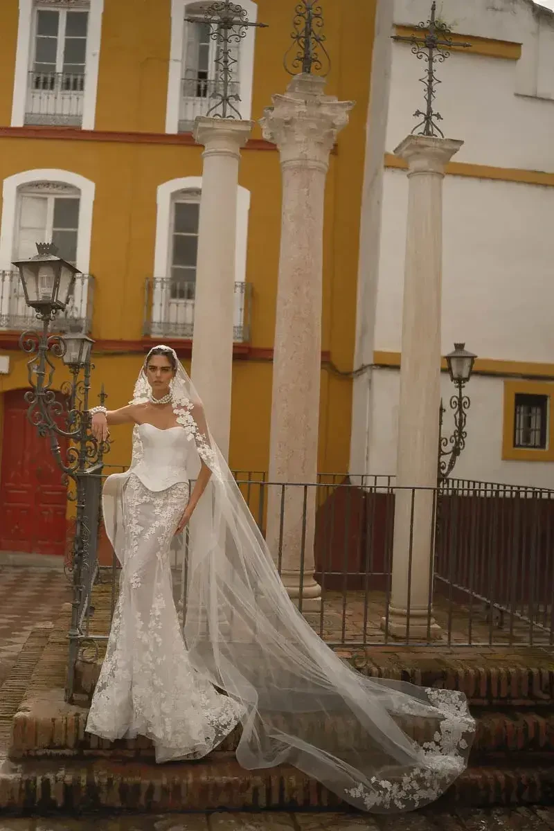 A bride in a white wedding gown with intricate lace details stands on steps, holding her veil and posing elegantly against a backdrop of columns and colorful architecture.