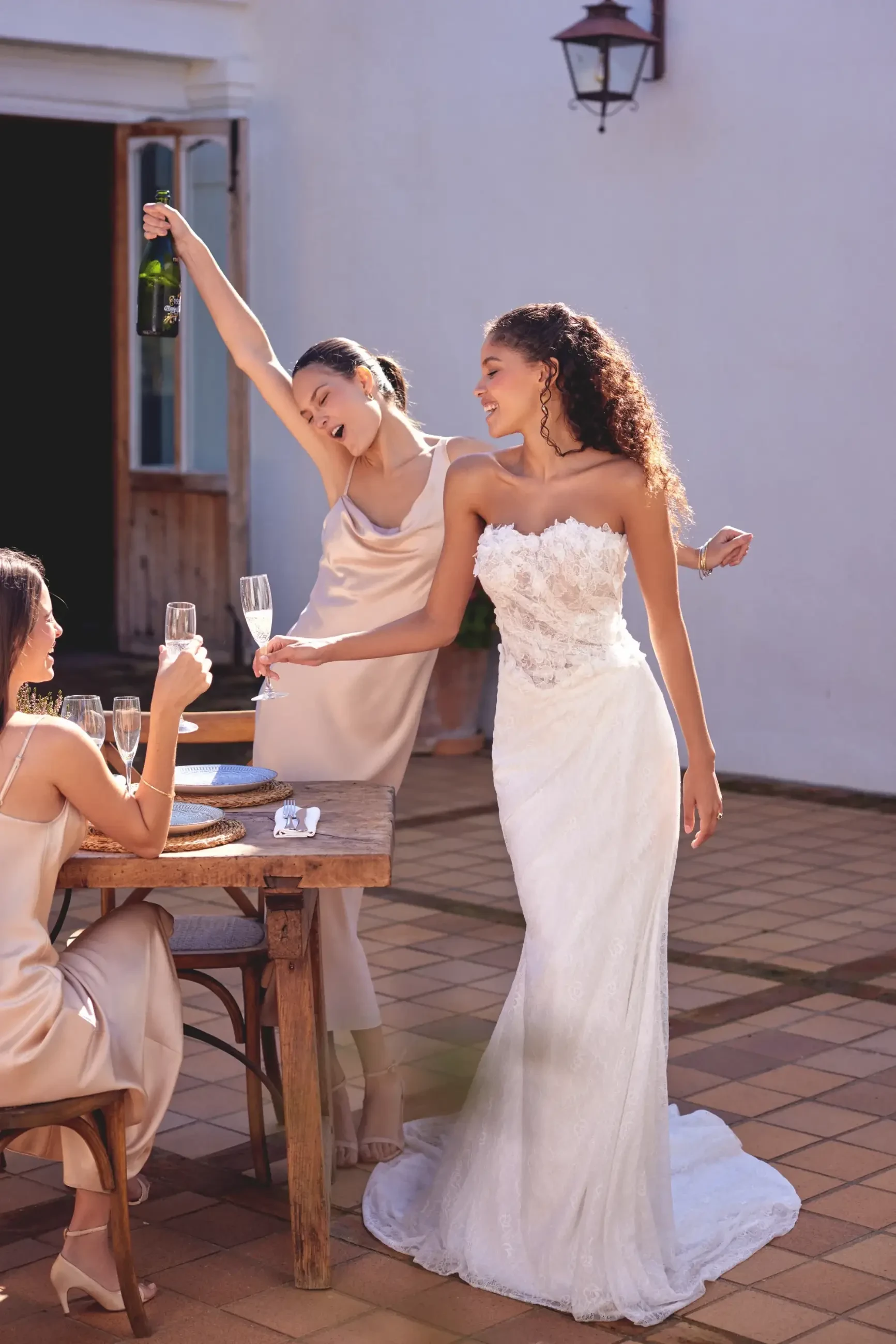 Bride in a lace gown smiles while holding a champagne glass. Two bridesmaids in satin dresses celebrate, one pouring champagne, on a sunlit patio.