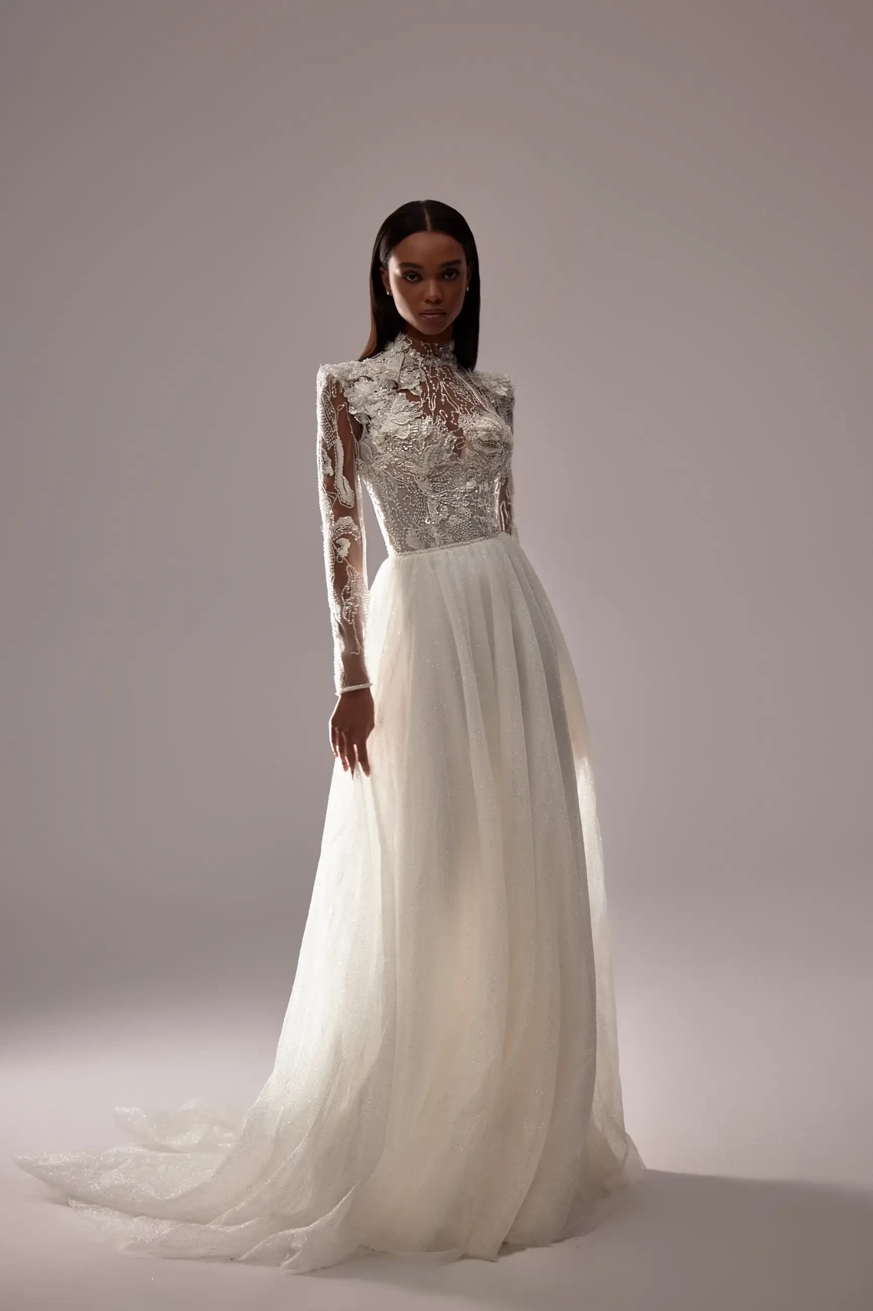 Elegant bridal gown with long lace sleeves, intricate bodice details, and flowing skirt. The model, set against a soft gray background, radiates sophistication.