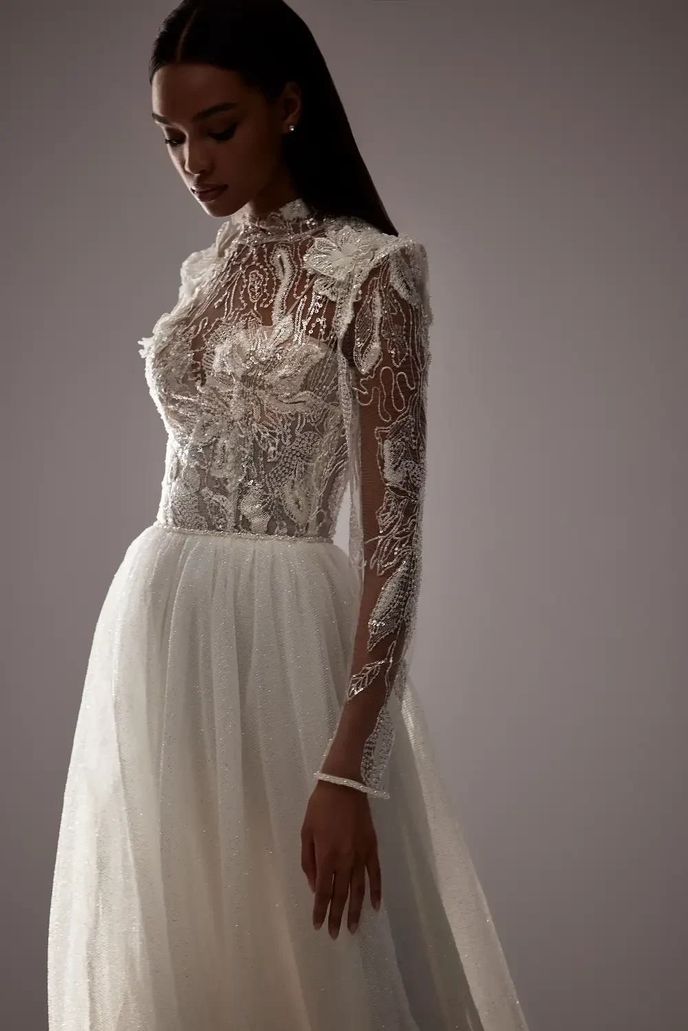 A model wearing an elegant, intricately designed white gown with sheer sleeves and beaded detailing.