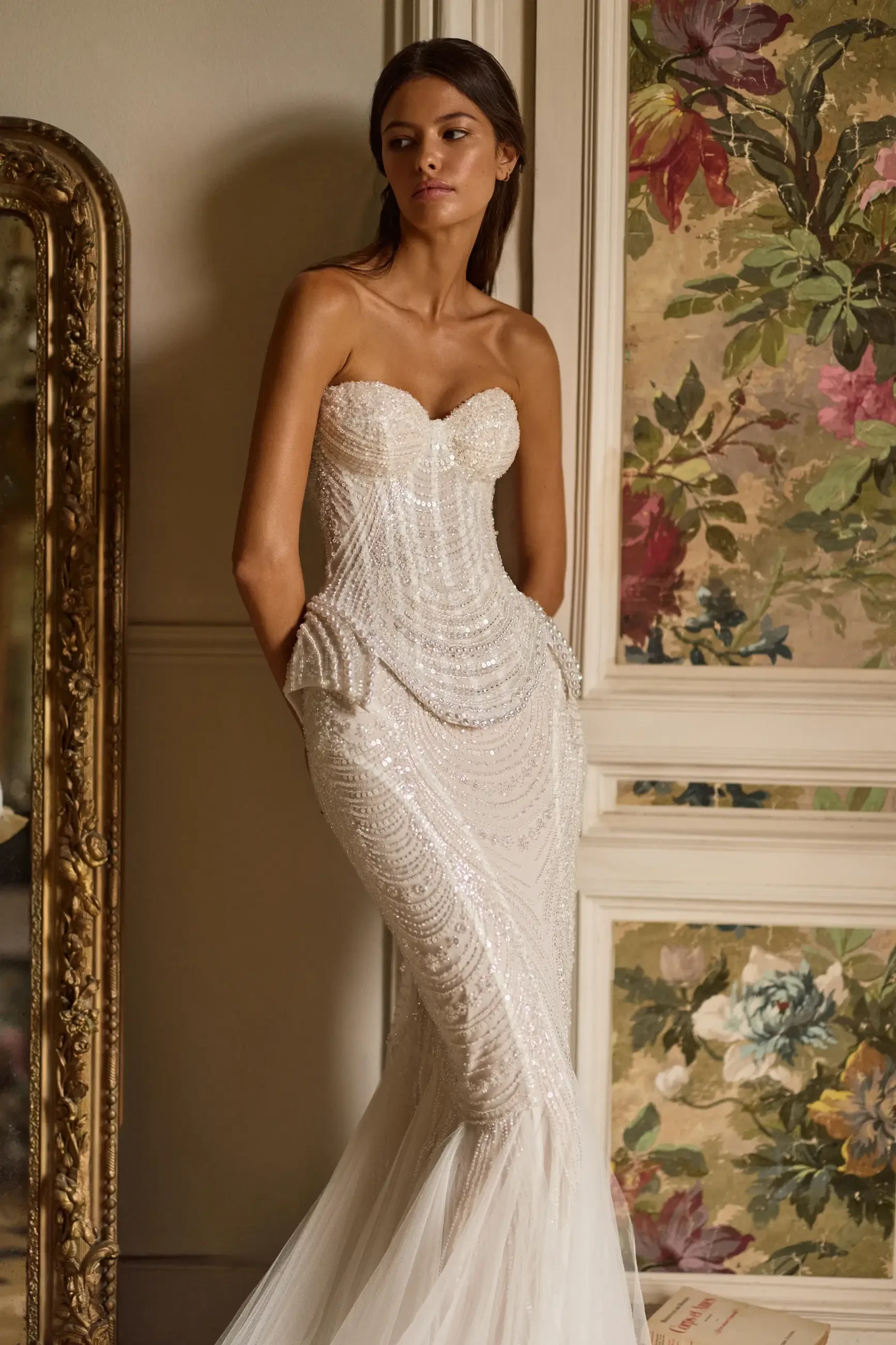 A model in a strapless, beaded white gown with a fitted silhouette, standing against a floral background and a vintage mirror.