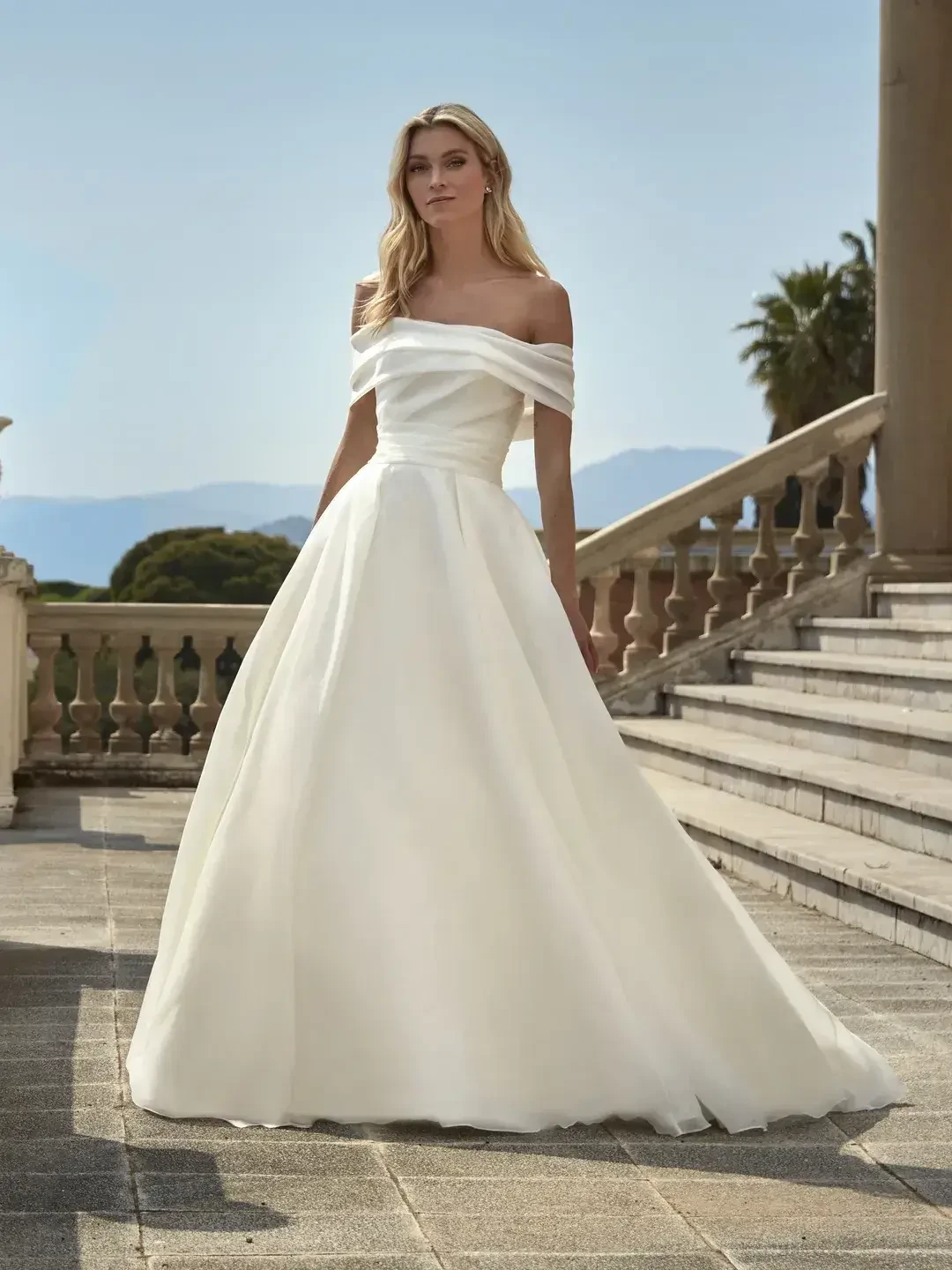 A bride in an elegant off-shoulder white gown stands on a stone terrace with steps. Behind her, clear skies and lush greenery create a serene backdrop.