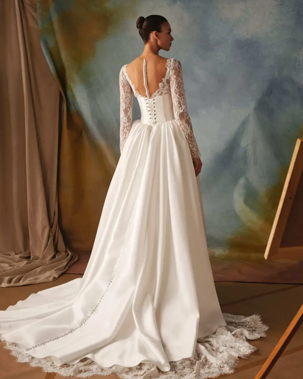 A woman is wearing a long-sleeved white wedding dress, standing with her back facing the camera. The dress features lace details and a flowing train, set against a softly colored background.