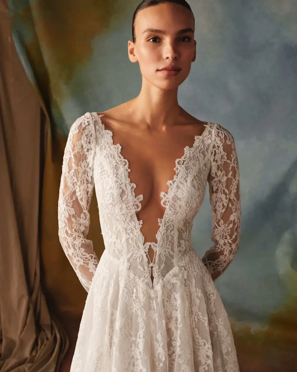 A woman wearing a long-sleeved, deep V-neck lace wedding dress, standing against a textured background.