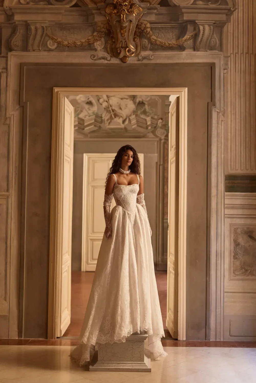 A woman in a white gown stands on a pedestal in a grand room with ornate walls and doors.