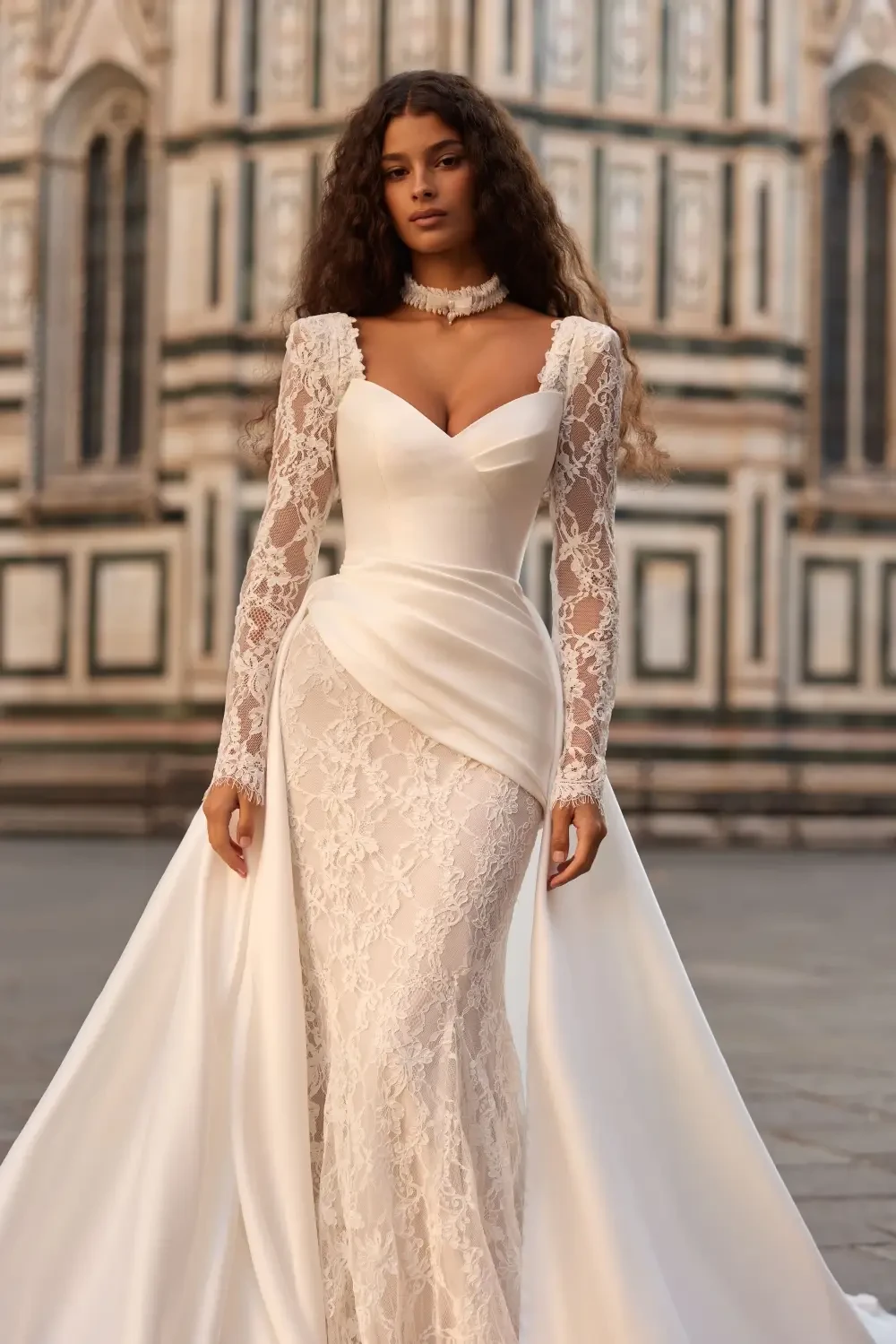 A model wearing an elegant white wedding dress with intricate lace details and long sleeves, standing in front of a beautifully ornate building.