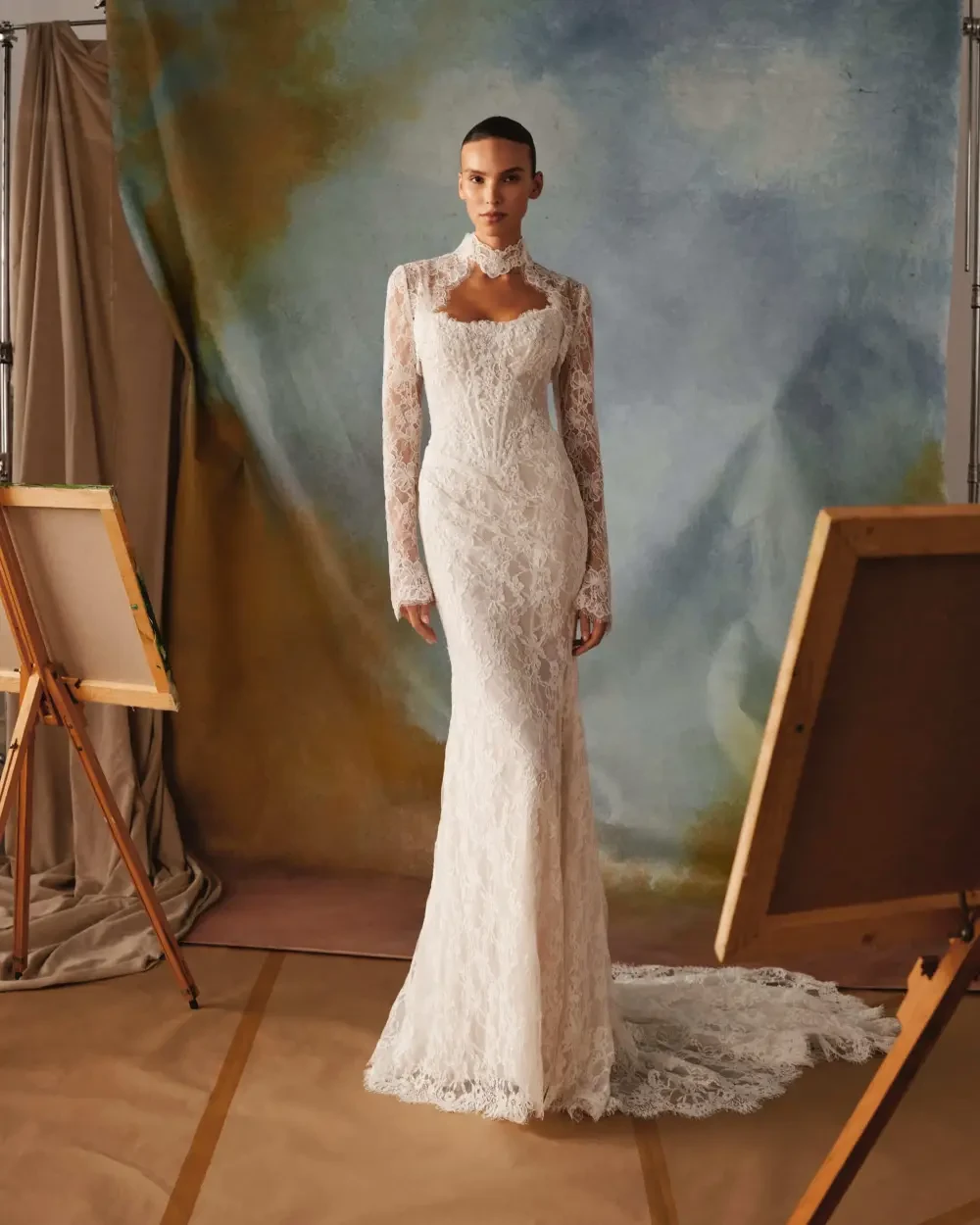 A model wearing a long-sleeved, lace wedding gown stands in front of a painted backdrop, with easels on either side.