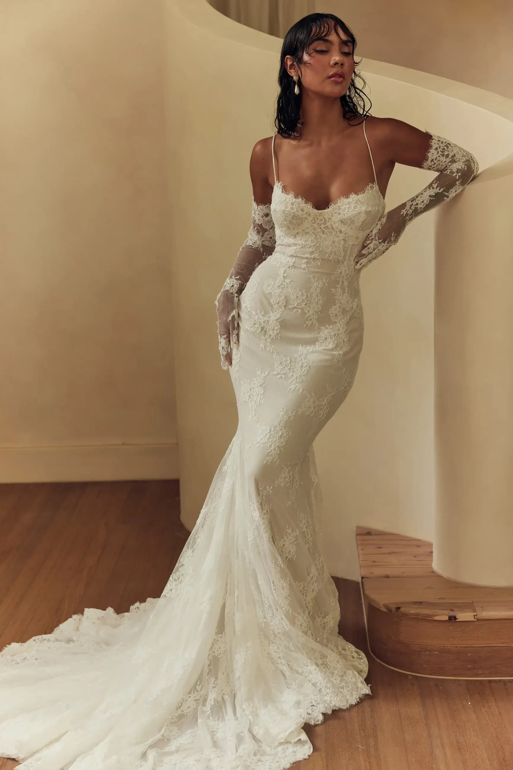 A model is wearing a fitted, lace wedding dress with a long train and thin straps, standing gracefully on a staircase. She has long hair and is accessorized with lace gloves.