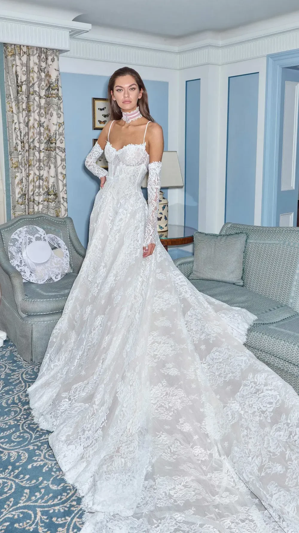 A model wearing an elegant white lace wedding gown, standing in a beautifully decorated room with vintage furniture and patterned curtains.