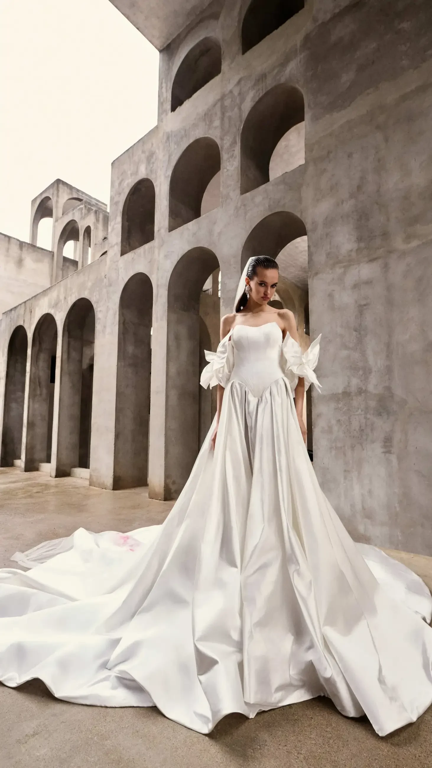 A woman stands in an elegant white gown with a long train, against a backdrop of tall, arched concrete structures. The scene conveys a sense of grandeur and sophistication.