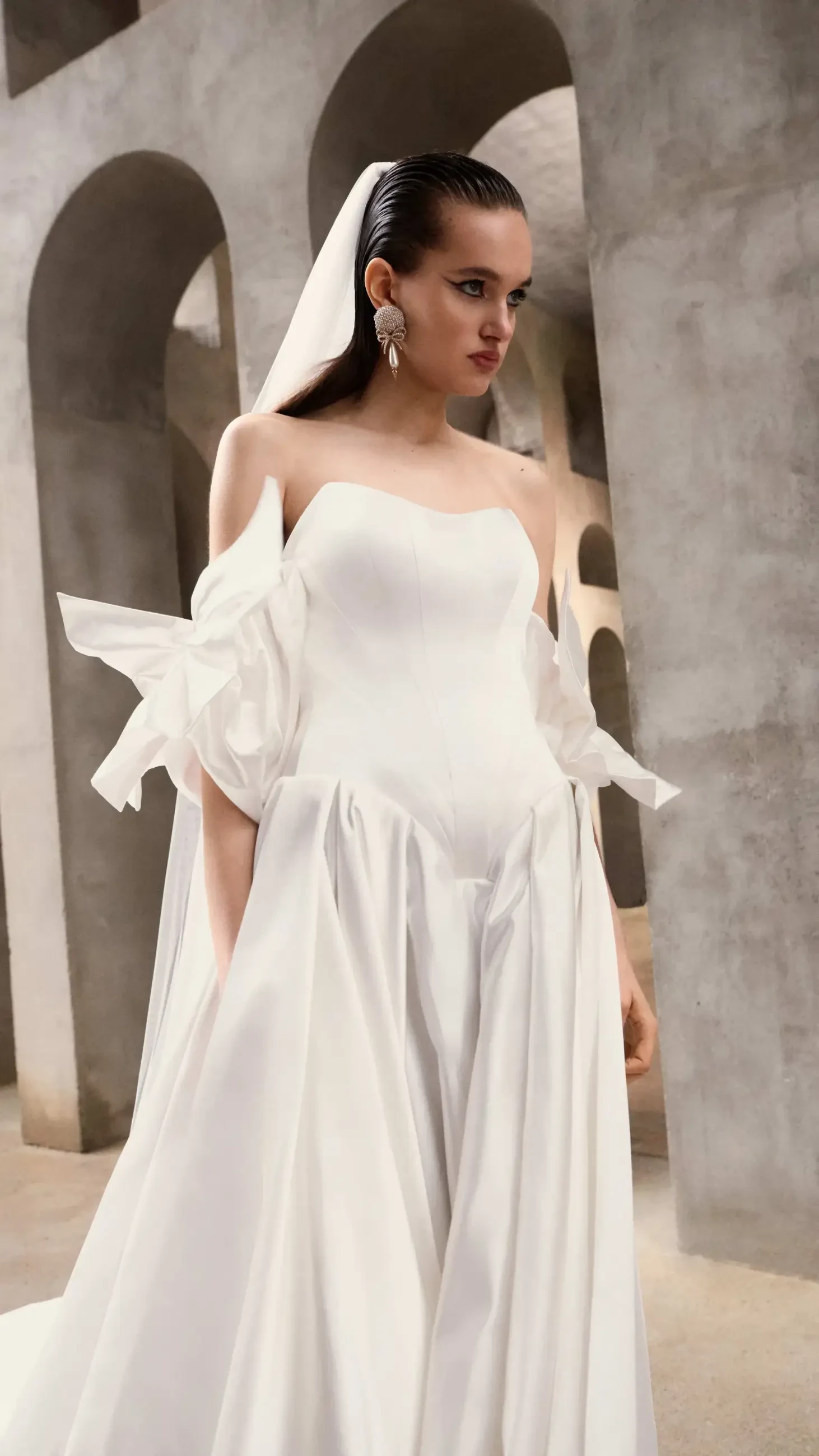 A model wearing a white wedding dress with dramatic sleeves, standing in an architectural space with arches.