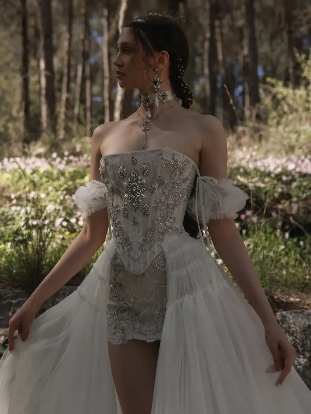 A woman in a strapless, embellished white dress with off-the-shoulder sleeves standing in a forested area with flowers in the background.