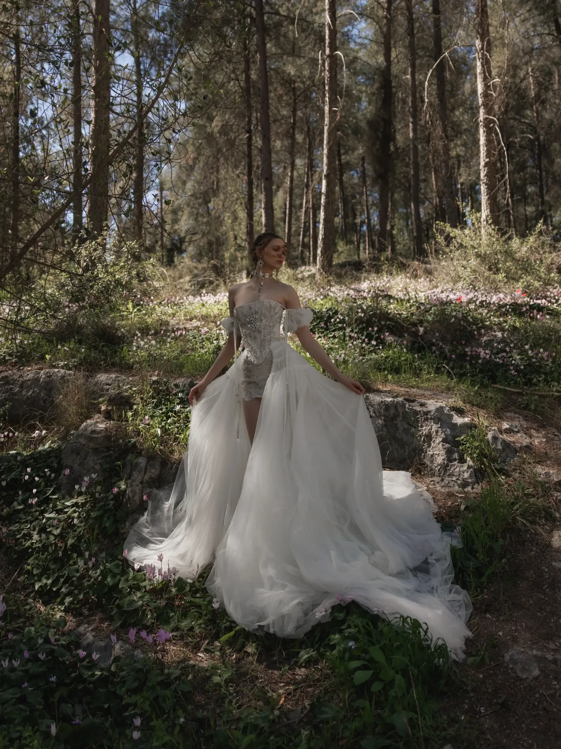 A woman in a flowing white dress stands in a forested area with trees and flowers around her.