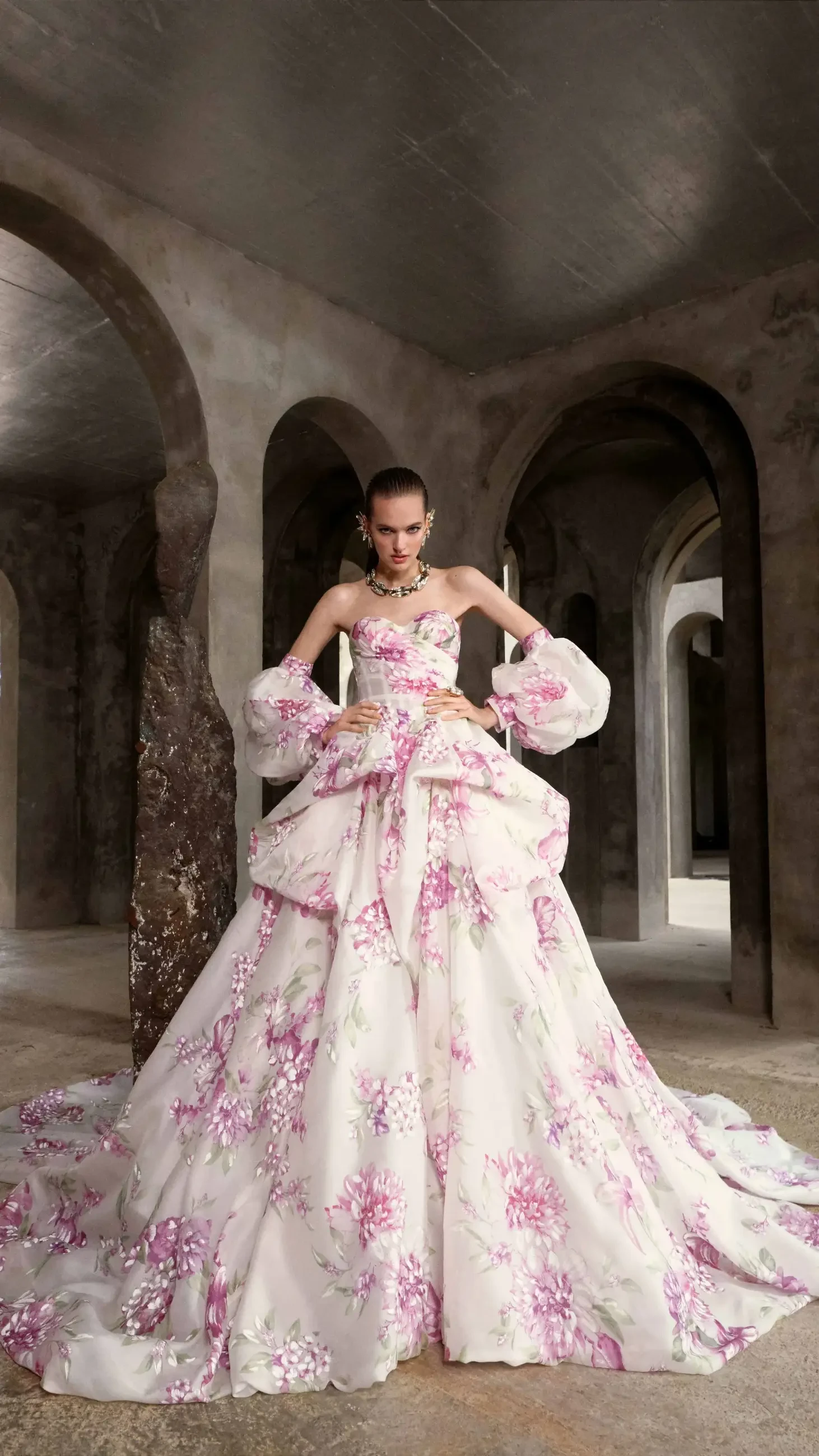 A woman in a floral gown stands in a stone hallway with arches, conveying elegance and confidence. The pink and white dress is ornate and voluminous.