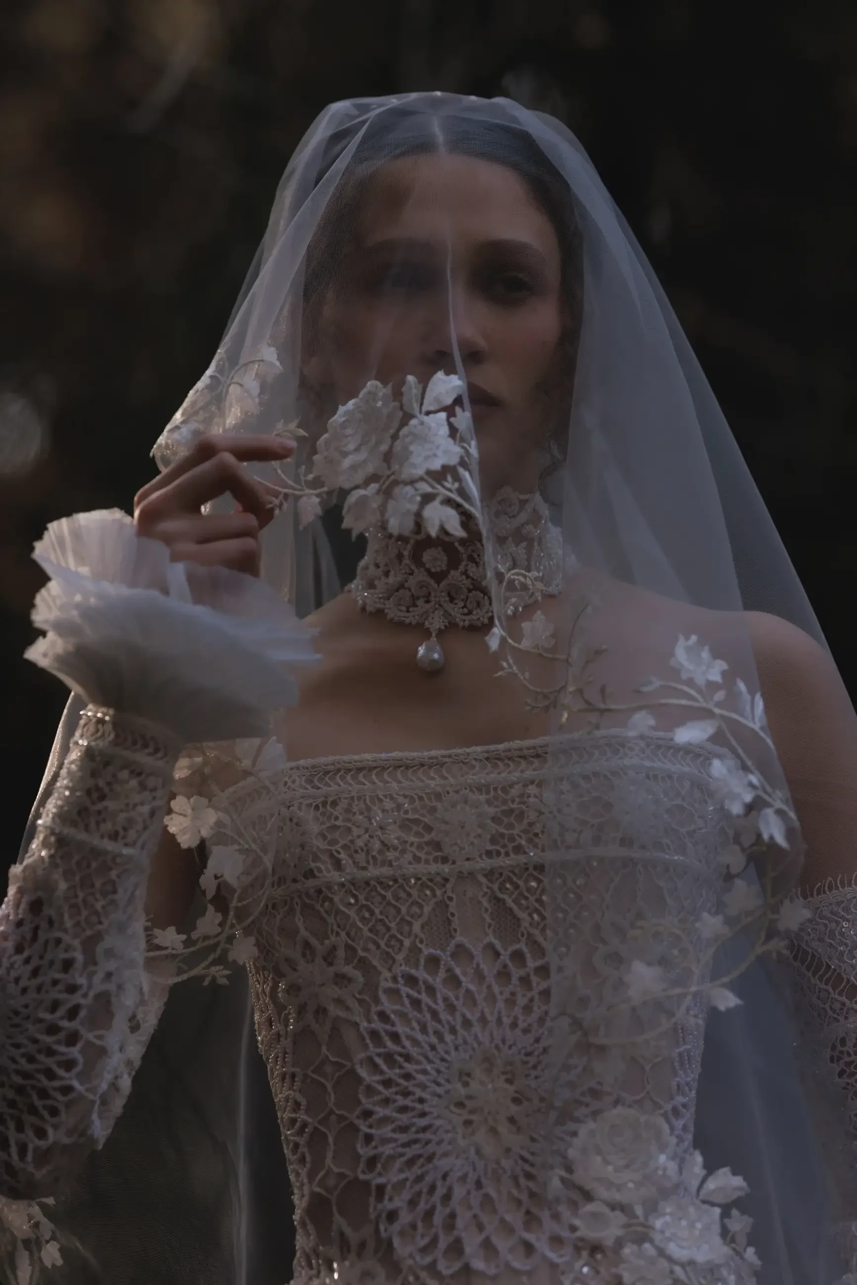 A woman in an intricate lace wedding dress with floral embellishments, holding a delicate lace handkerchief and looking directly at the camera, partially obscured by a sheer veil.
