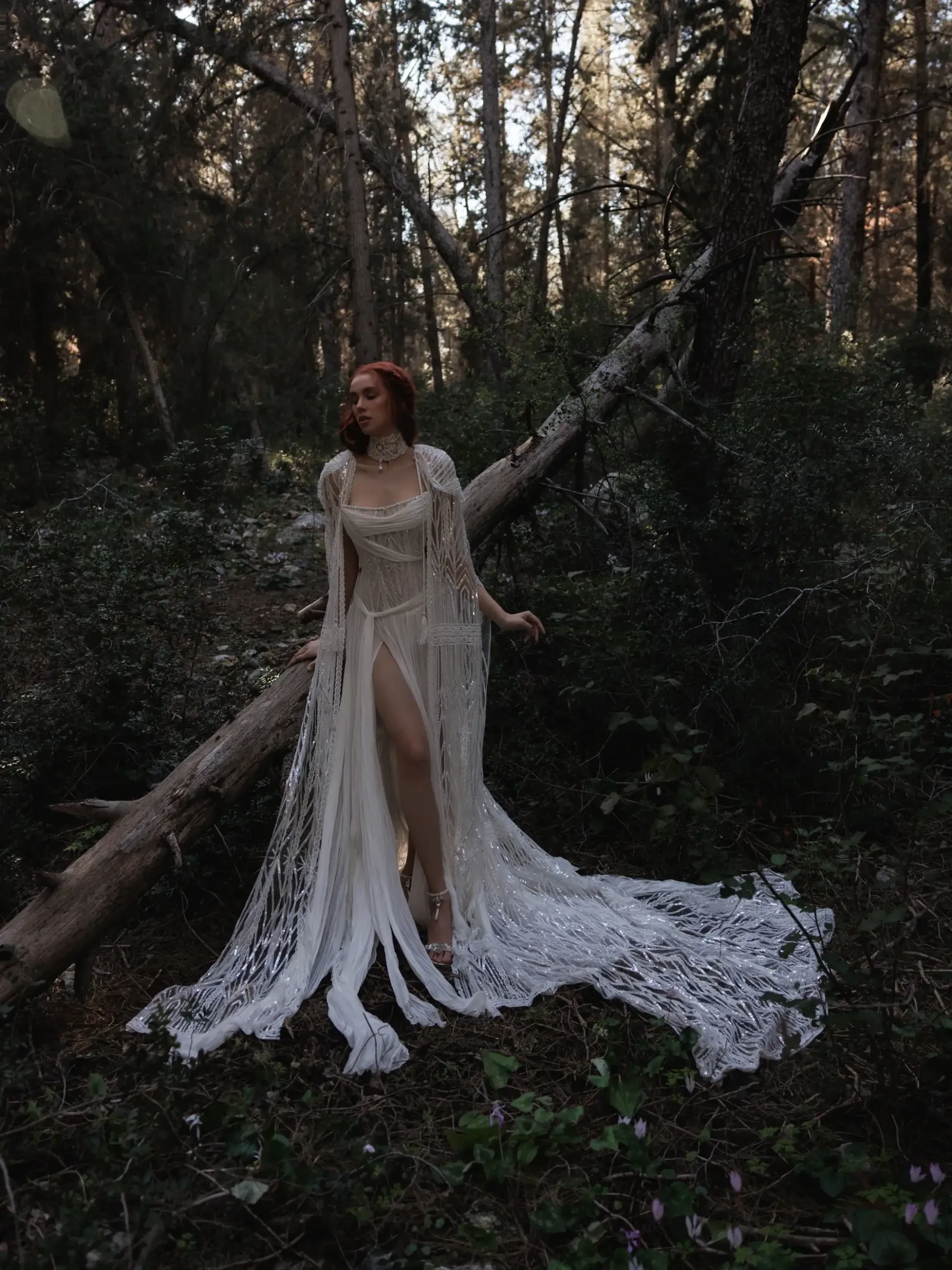 A woman stands in a wooded area, wearing a flowing white garment with fringes. She poses thoughtfully near a fallen tree, surrounded by greenery and soft light filtering through the trees.