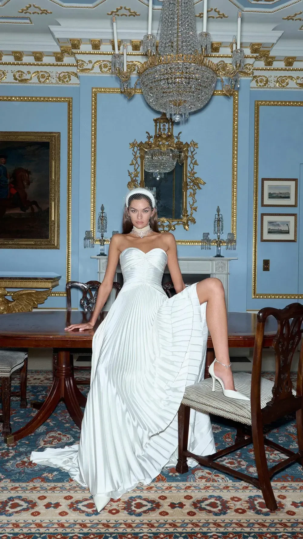 A model in a white strapless gown poses elegantly in a lavishly decorated room with ornate blue walls and a chandelier.