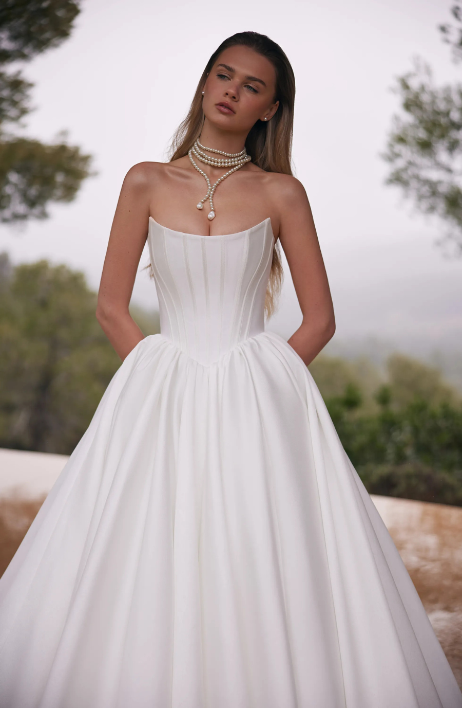 A woman in a strapless white wedding gown with a full skirt stands outdoors. She wears a layered pearl necklace. The setting is serene and elegant.