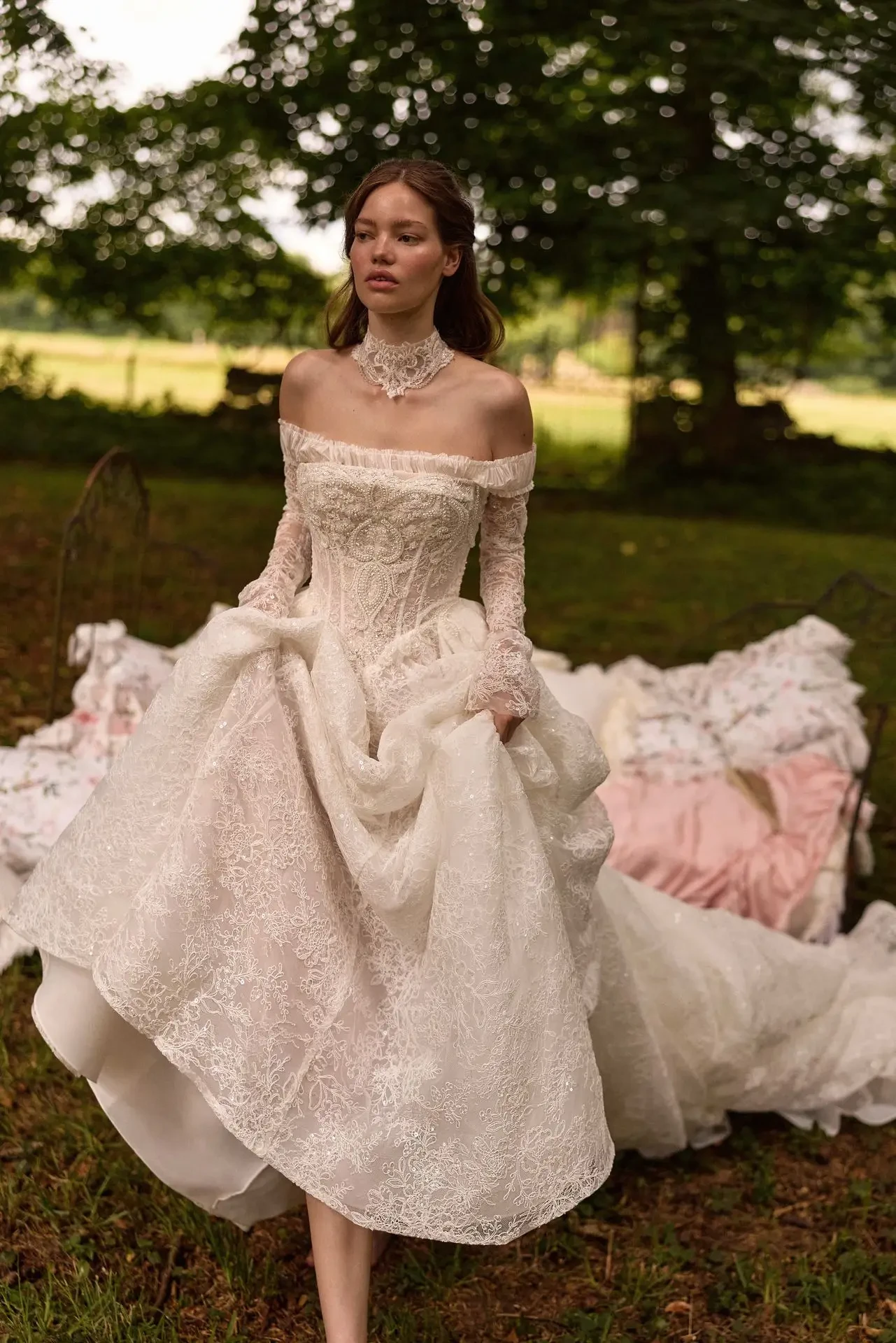 A woman in an elegant off-shoulder lace wedding gown stands outdoors, surrounded by greenery. She looks serene, holding her dress as sunlight filters through the trees.