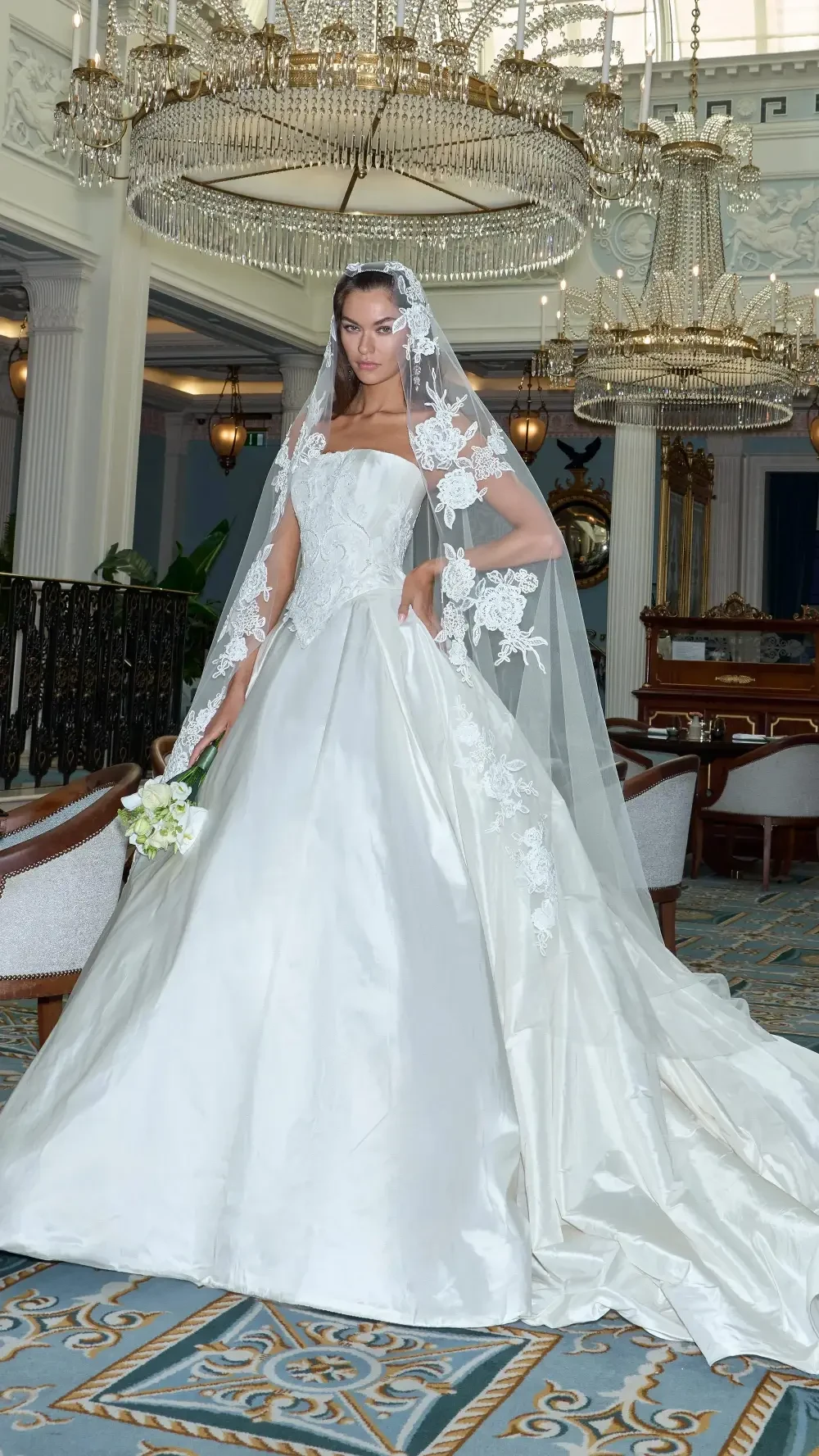 A model wearing a stunning white wedding gown with lace details and a long veil, standing in an elegant indoor setting with chandeliers.