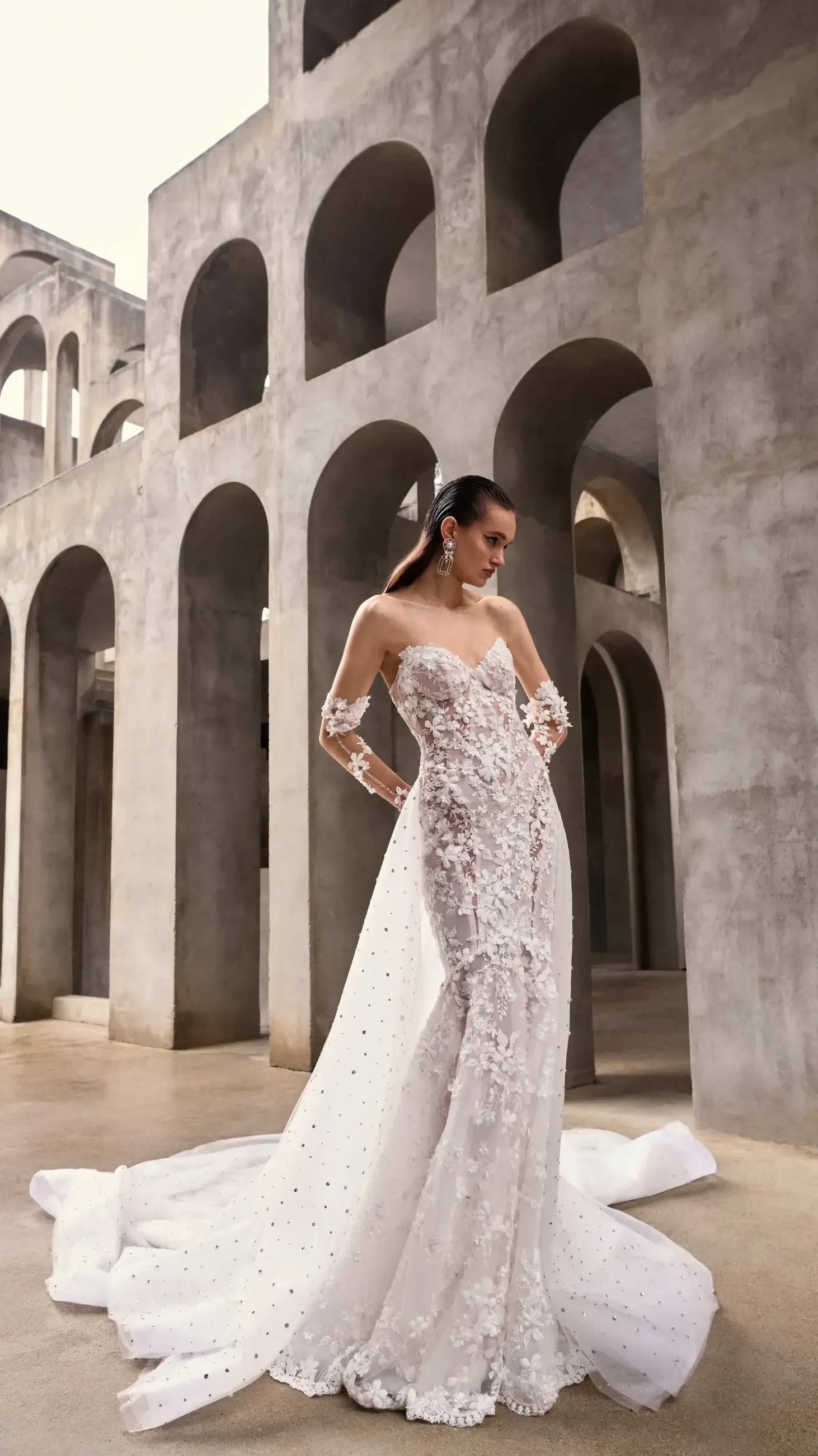 A model wearing a stunning wedding dress with intricate beadwork and a long train, standing in an architectural space with arches.