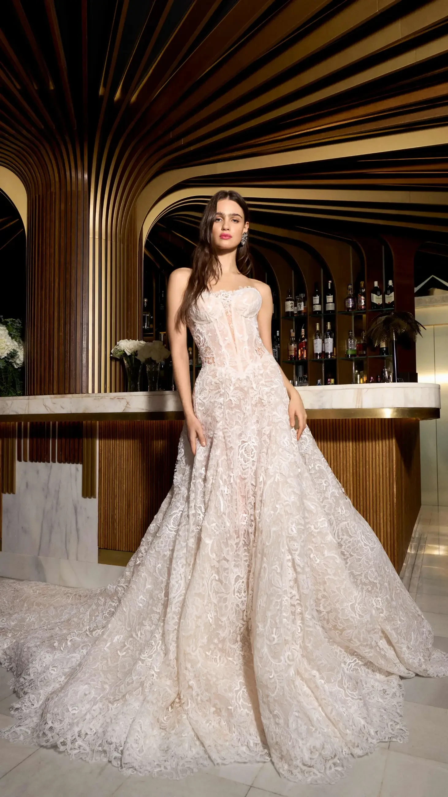 A woman in an elegant, lace wedding gown stands confidently in a stylish bar setting with gold and wood accents, exuding sophistication and grace.