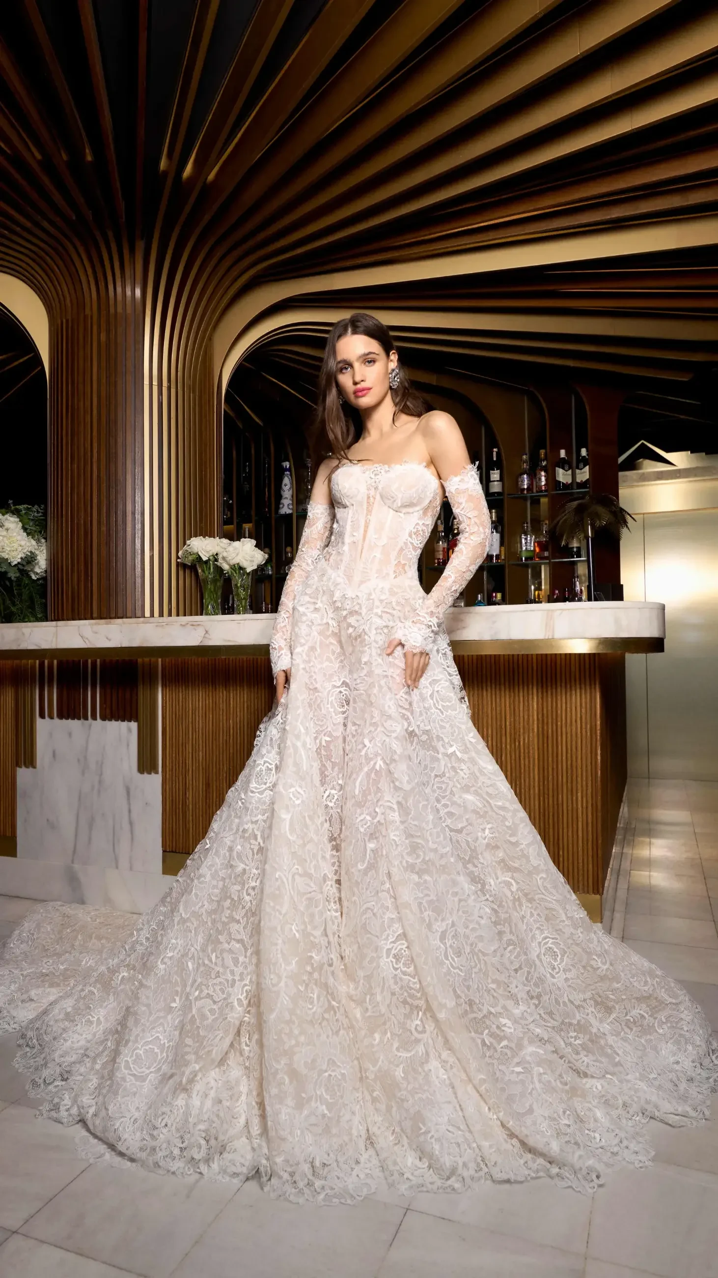 A woman in an elegant, lace-covered wedding gown stands confidently in a stylish bar with a marble counter. The ambiance is luxurious and sophisticated.