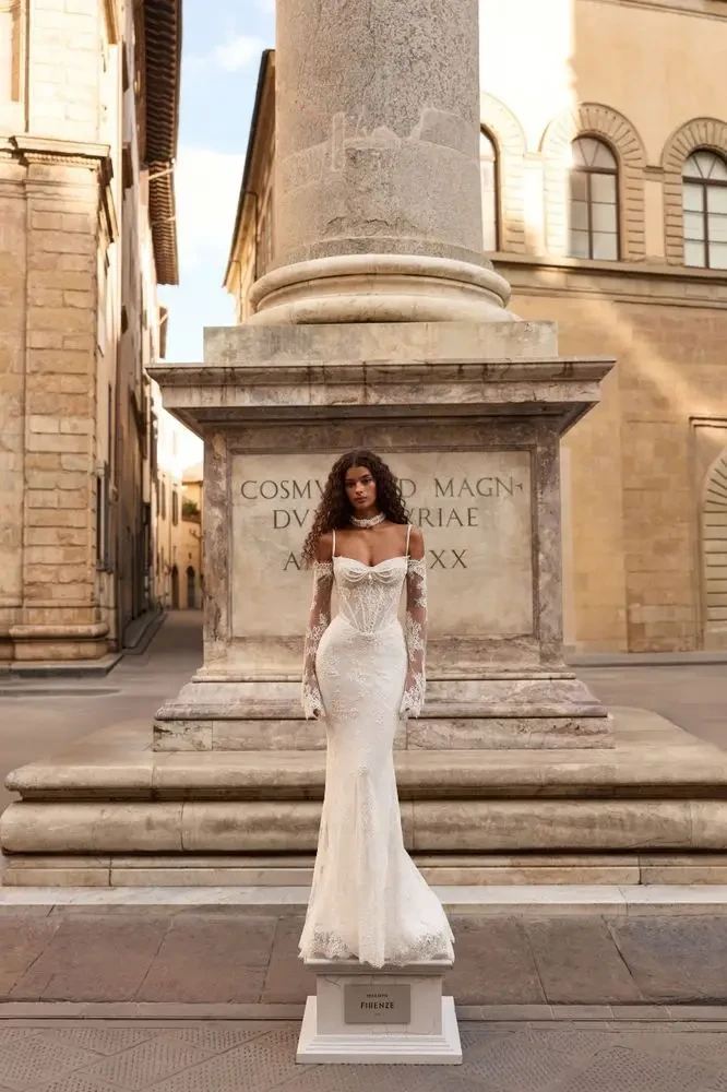 A woman in an elegant white dress stands on a pedestal in a historic street setting, with an old stone column and buildings in the background.