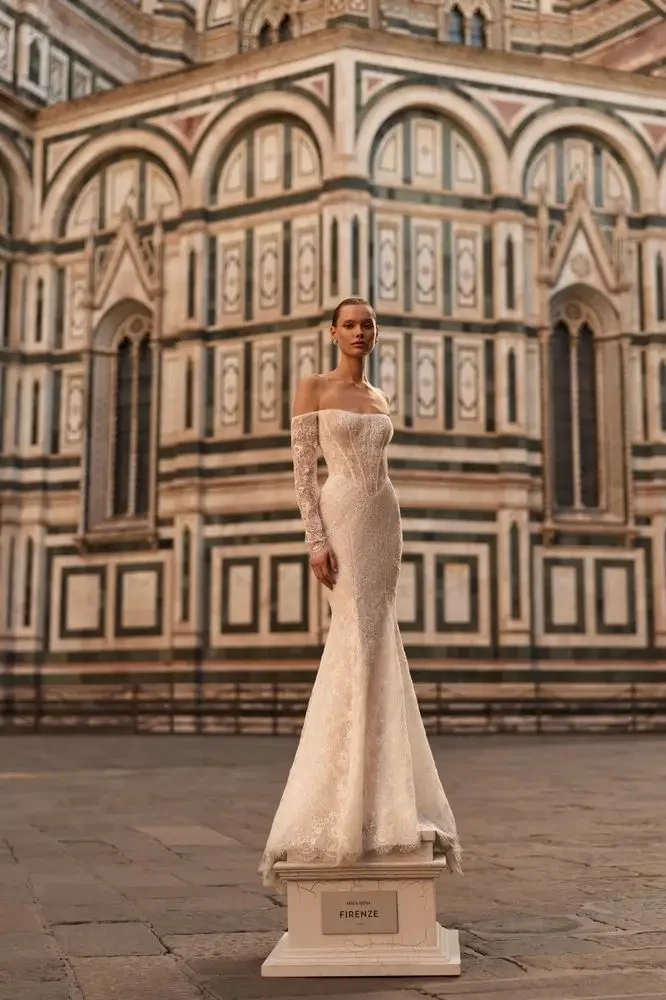 A model wearing a white off-the-shoulder wedding dress poses in front of a historical building with intricate architectural details.