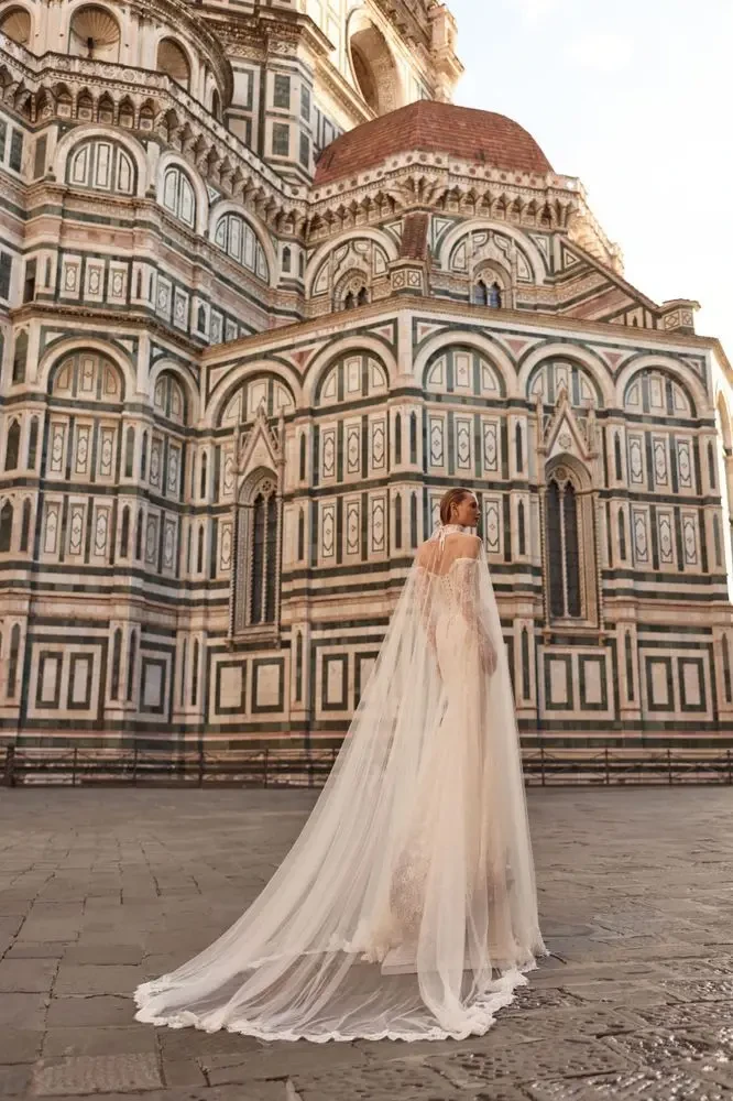 A model wearing a white off-the-shoulder wedding dress poses in front of a historical building with intricate architectural details.