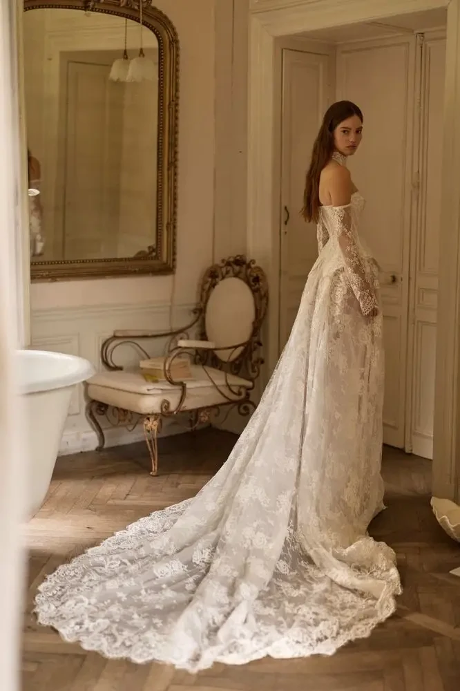 A woman in a white lace wedding dress stands in a vintage-style room, facing away from the camera. The dress features a long train and off-the-shoulder sleeves, and there is a decorative mirror and a vintage chair in the background.