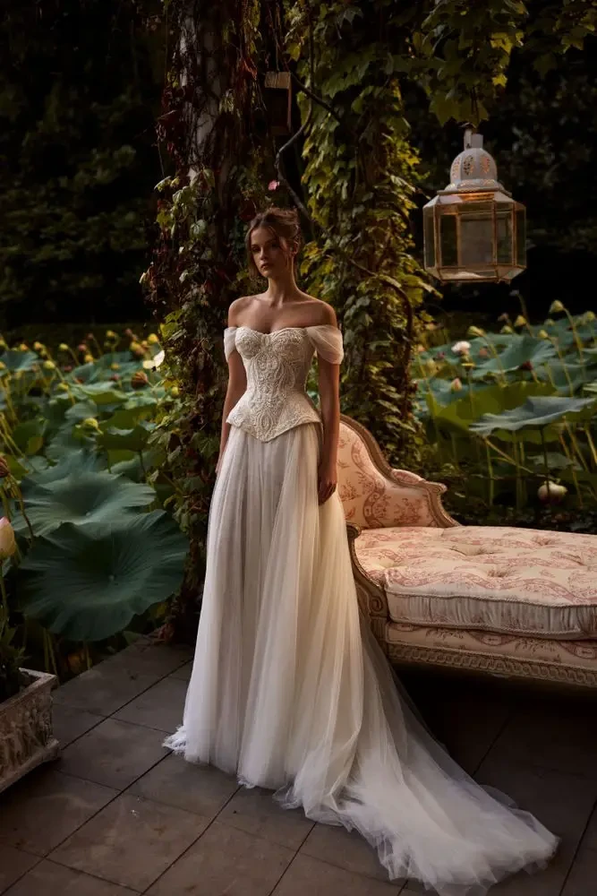 A woman in an elegant off-the-shoulder wedding dress stands in a garden with lush greenery and lotus flowers, next to a vintage sofa.