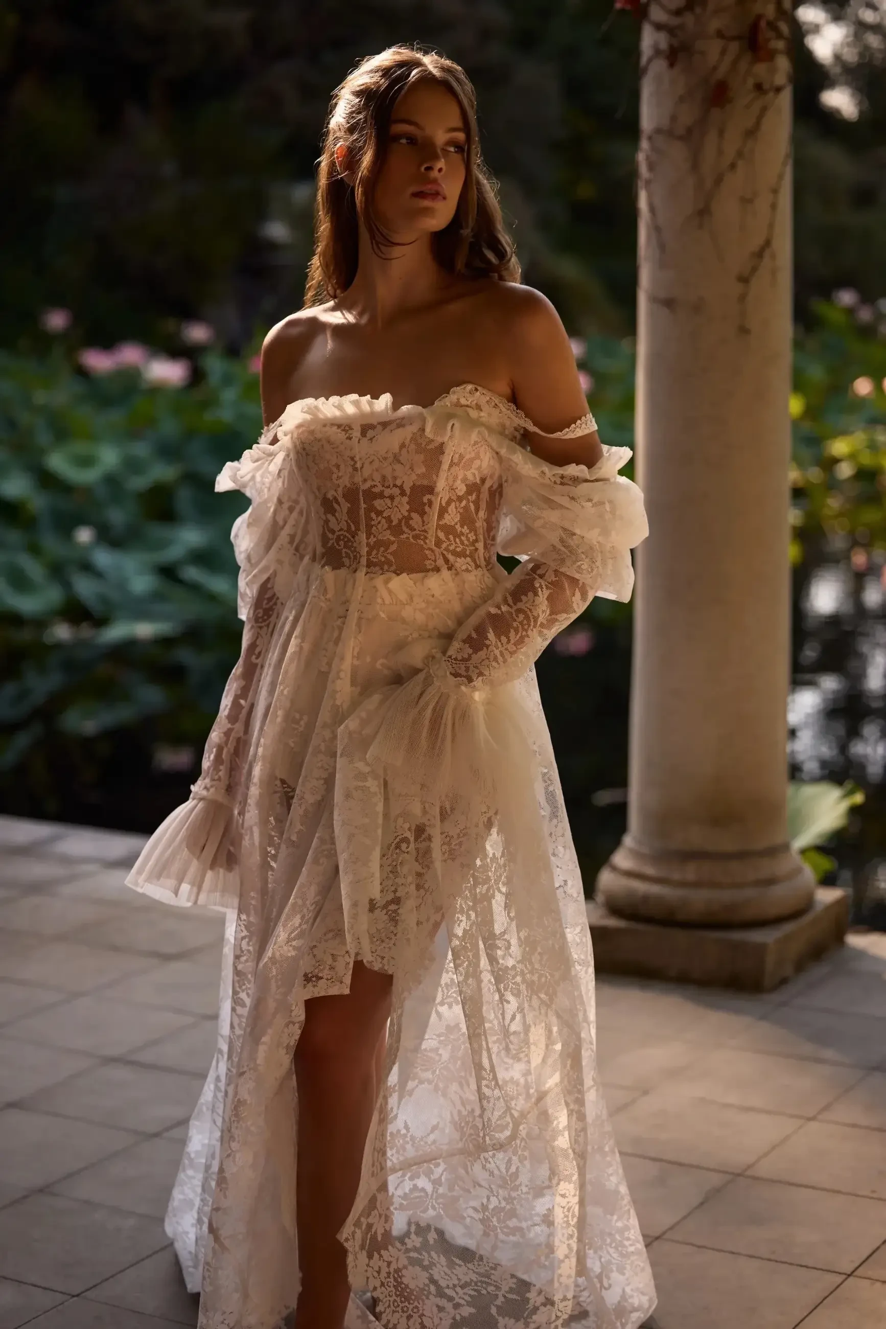 A woman in a delicate, off-the-shoulder lace gown stands by a column, bathed in warm sunlight. She gazes thoughtfully, surrounded by lush greenery.