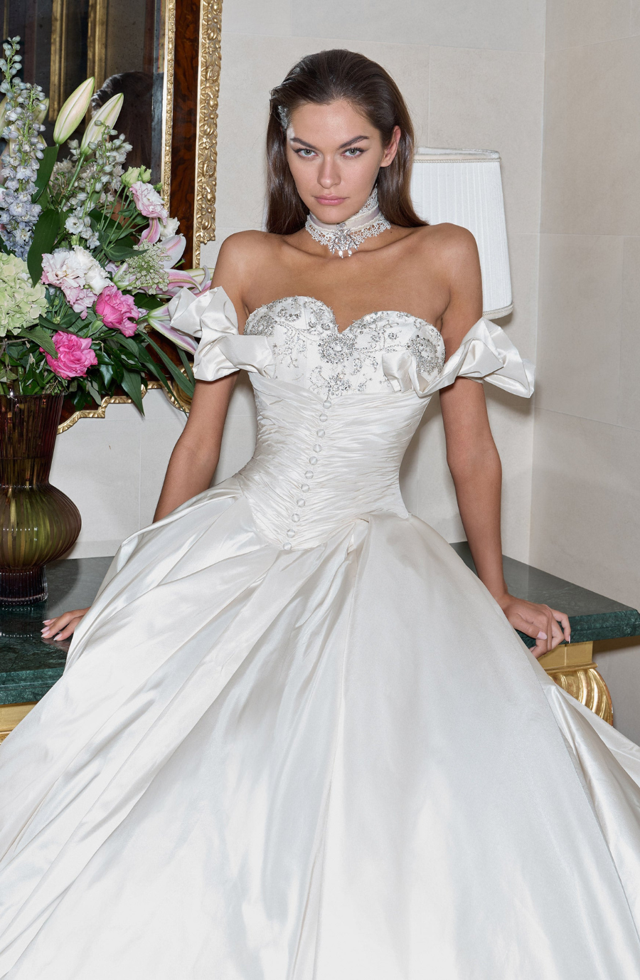 A woman in an elegant white wedding gown with embellishments poses confidently. She wears a sparkling choker. A bouquet of colorful flowers is in the background.