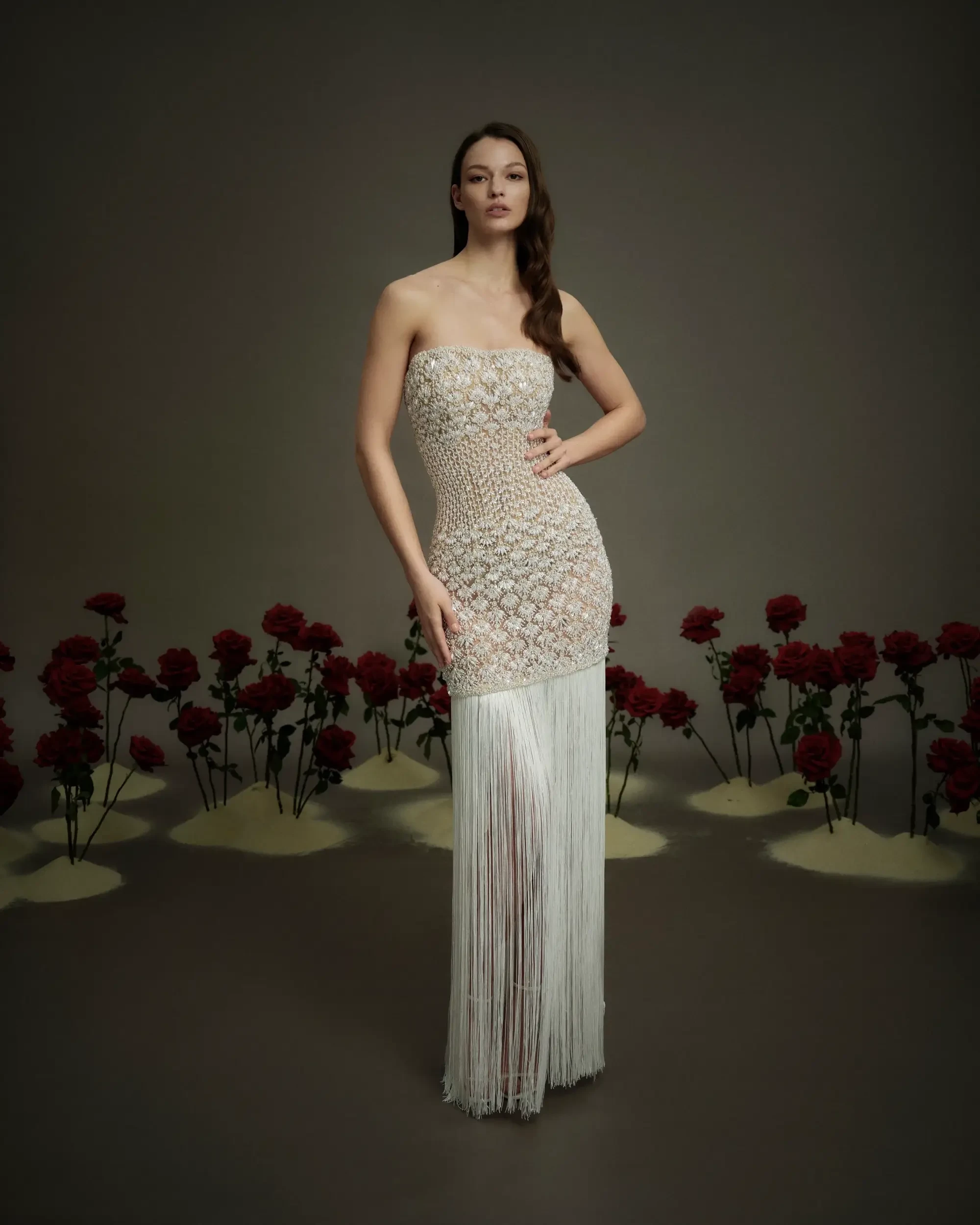 A model wearing a strapless beaded gown with fringe, standing in front of a backdrop of red roses and sand.