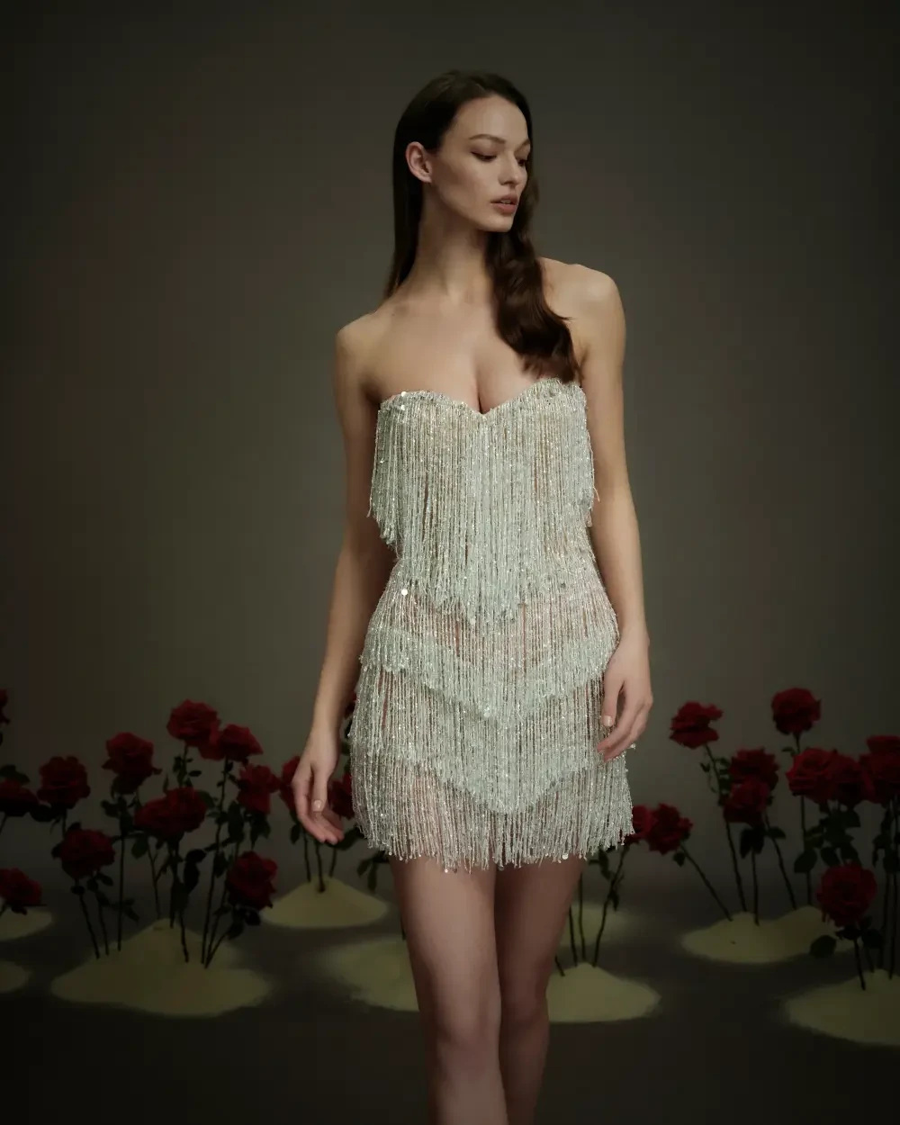 A model wearing a shimmering, crystal-embellished dress, standing against a dark backdrop with red roses positioned in the foreground.