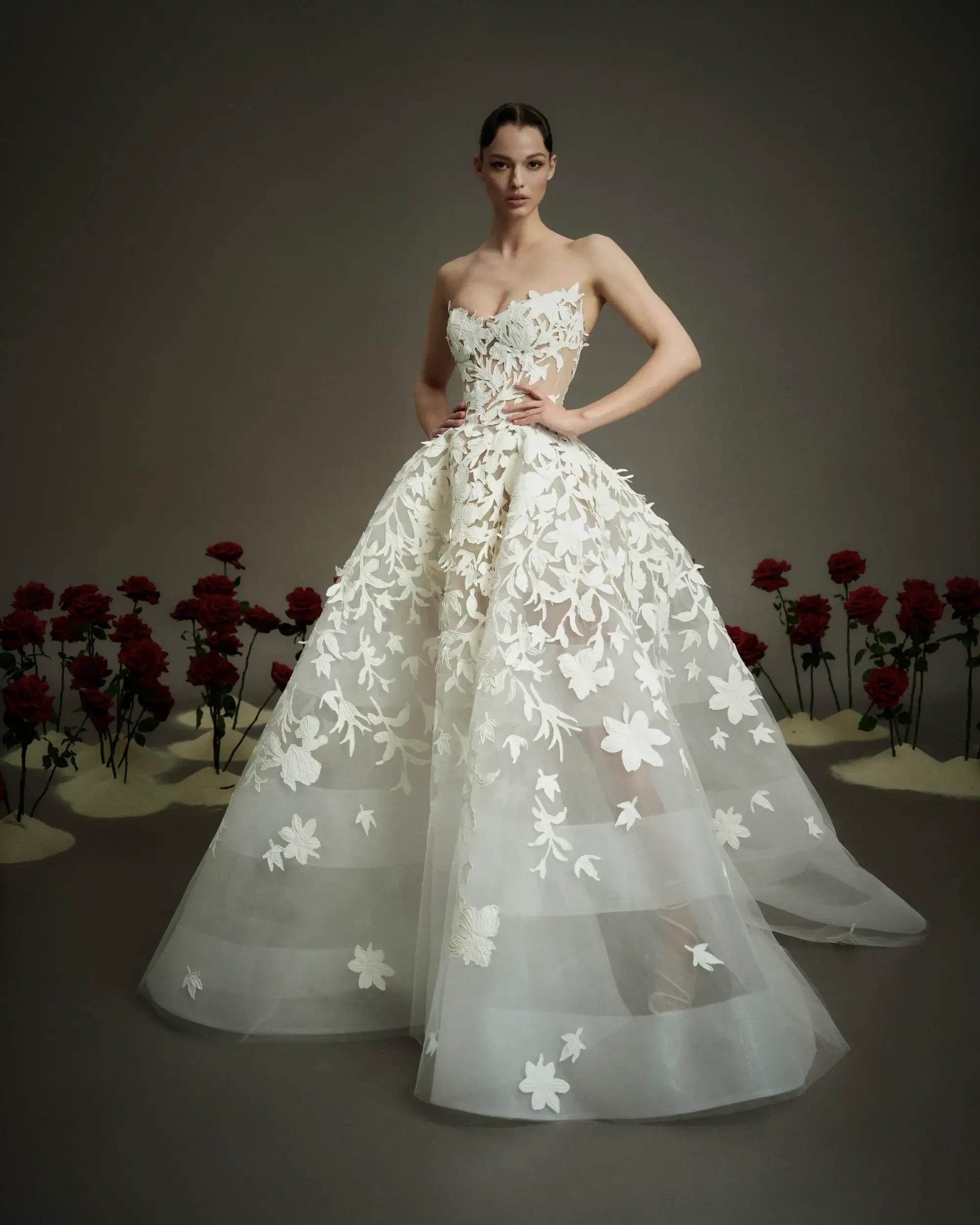 A model wearing a strapless gown adorned with floral appliqu&eacute;s, standing in front of roses against a neutral background.