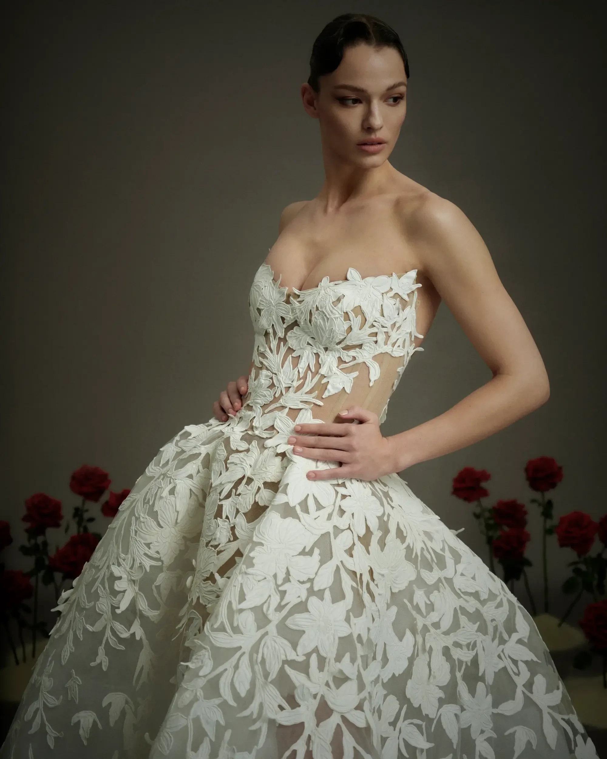 A model poses in a strapless white dress adorned with floral cutouts, against a muted background with red roses.