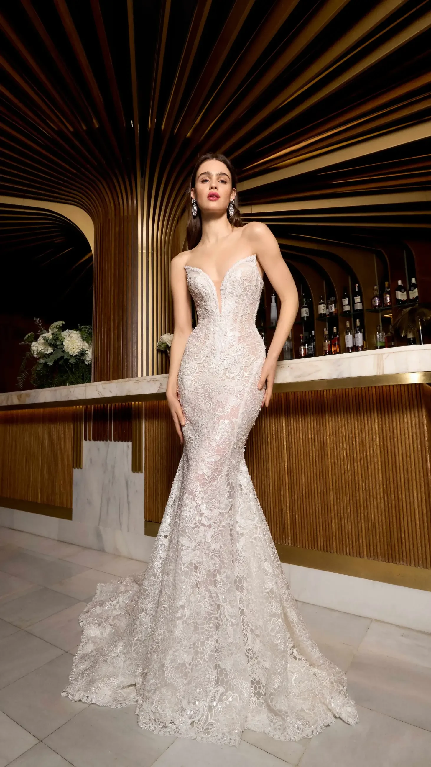 A woman wearing a form-fitting, lace gown with a sweetheart neckline poses confidently in front of an elegant bar. The background features wooden paneling and soft lighting, enhancing the luxurious atmosphere.