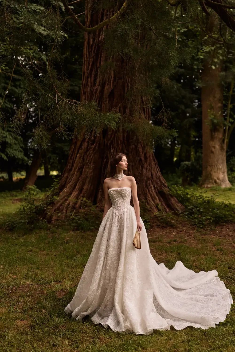 A woman in a strapless, elegant wedding gown stands gracefully in a lush green setting, with a large tree in the background.