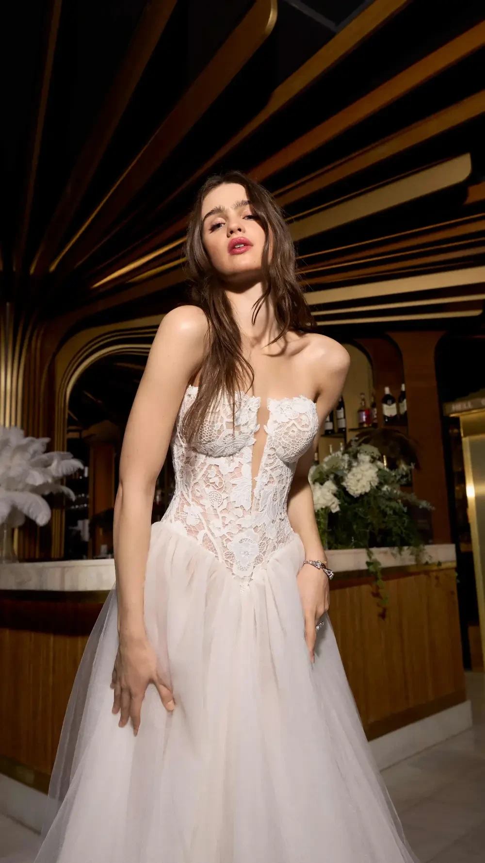 A woman in a white, elegant dress with a sheer overlay poses in an upscale setting with stylish decor in the background.