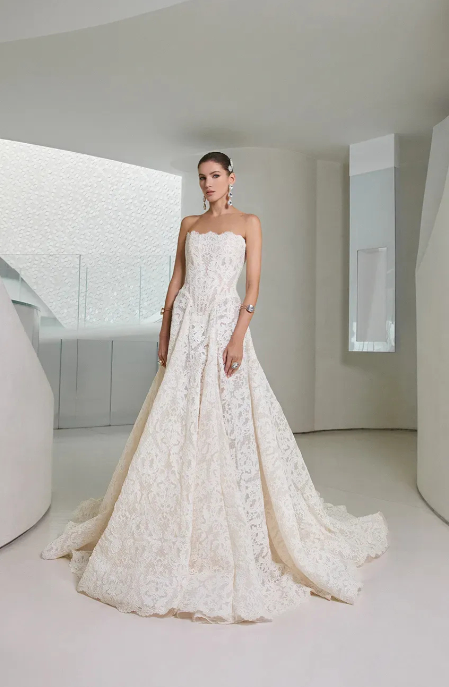 A woman stands elegantly in a strapless lace wedding dress with a long train in a modern, white, minimalist room, exuding a sophisticated, serene aura.