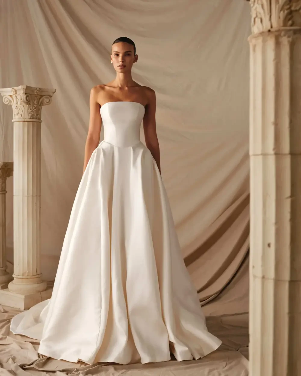 A model wearing a strapless white gown stands in front of a neutral-toned backdrop with columns.